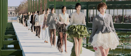 Desfile ao ar livre em Paris: fila de modelos na passarela branca entre estruturas verdes e espelho d'água com vitórias-régias