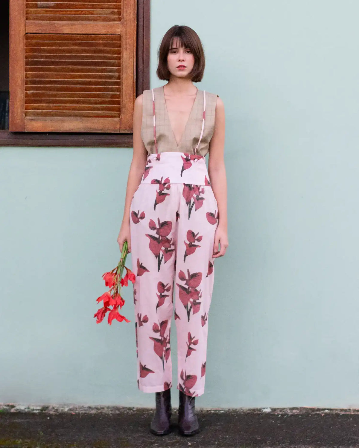 Modelo com colete bege e calça clara estampada com flores vermelhas, segurando um buquê de flores vermelhas, encostada em uma parede verde-água ao lado de uma janela de madeira